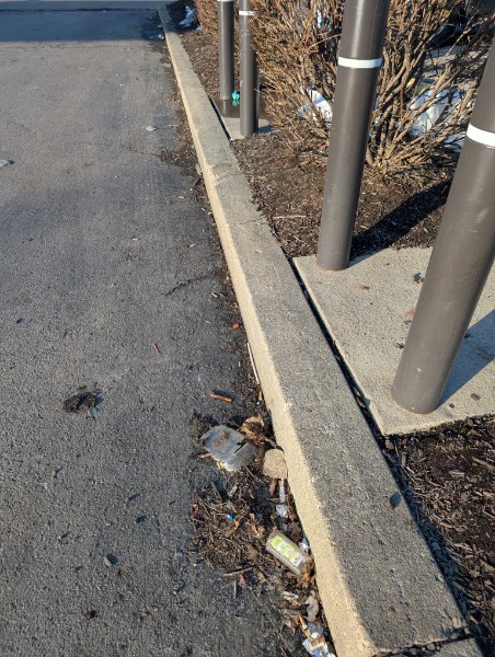 Parking lot debris and litter gathered along a curb beside bollards at a commercial site